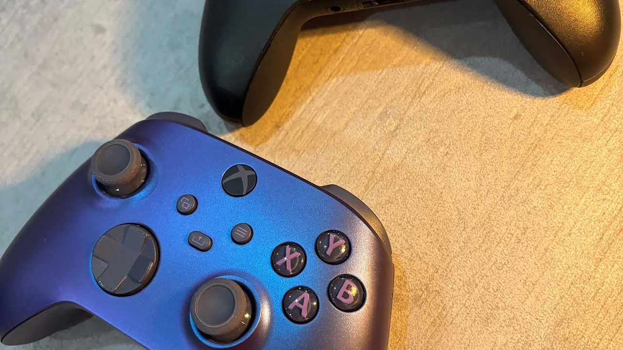 Two Xbox game controllers lie next to each other on a light, subtly textured surface. The top controller is black, while the bottom one has a blue-to-purple gradient finish. Both feature two analog sticks, a directional pad, menu and function buttons, and four colored action buttons labeled A, B, X, and Y. Warm light from the right casts soft shadows and highlights the surface texture. — cover image for Xbox Brings Four Backward-Compatible Games to PC and Handhelds