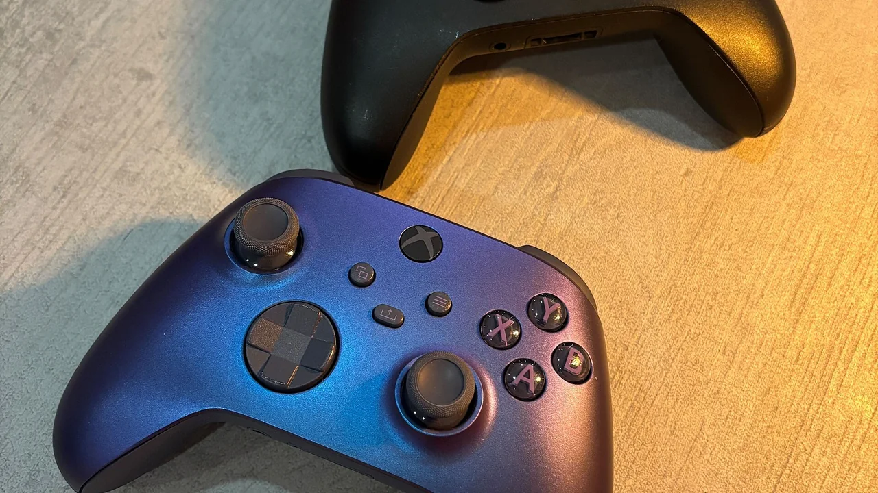 Two Xbox game controllers lie next to each other on a light, subtly textured surface. The top controller is black, while the bottom one has a blue-to-purple gradient finish. Both feature two analog sticks, a directional pad, menu and function buttons, and four colored action buttons labeled A, B, X, and Y. Warm light from the right casts soft shadows and highlights the surface texture. — cover image for Xbox Mode Begins Rolling Out to Windows 11 PCs